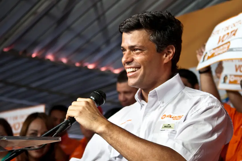 Leopoldo López smiles while speaking into a microphone at a Voluntad Popular event, supporters holding signs behind him.